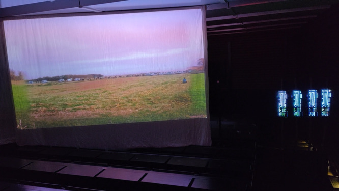 A videobeam-screen showing overlayed image of police line on meadow. 4 keyboard installition beside the screen. in a dark room.